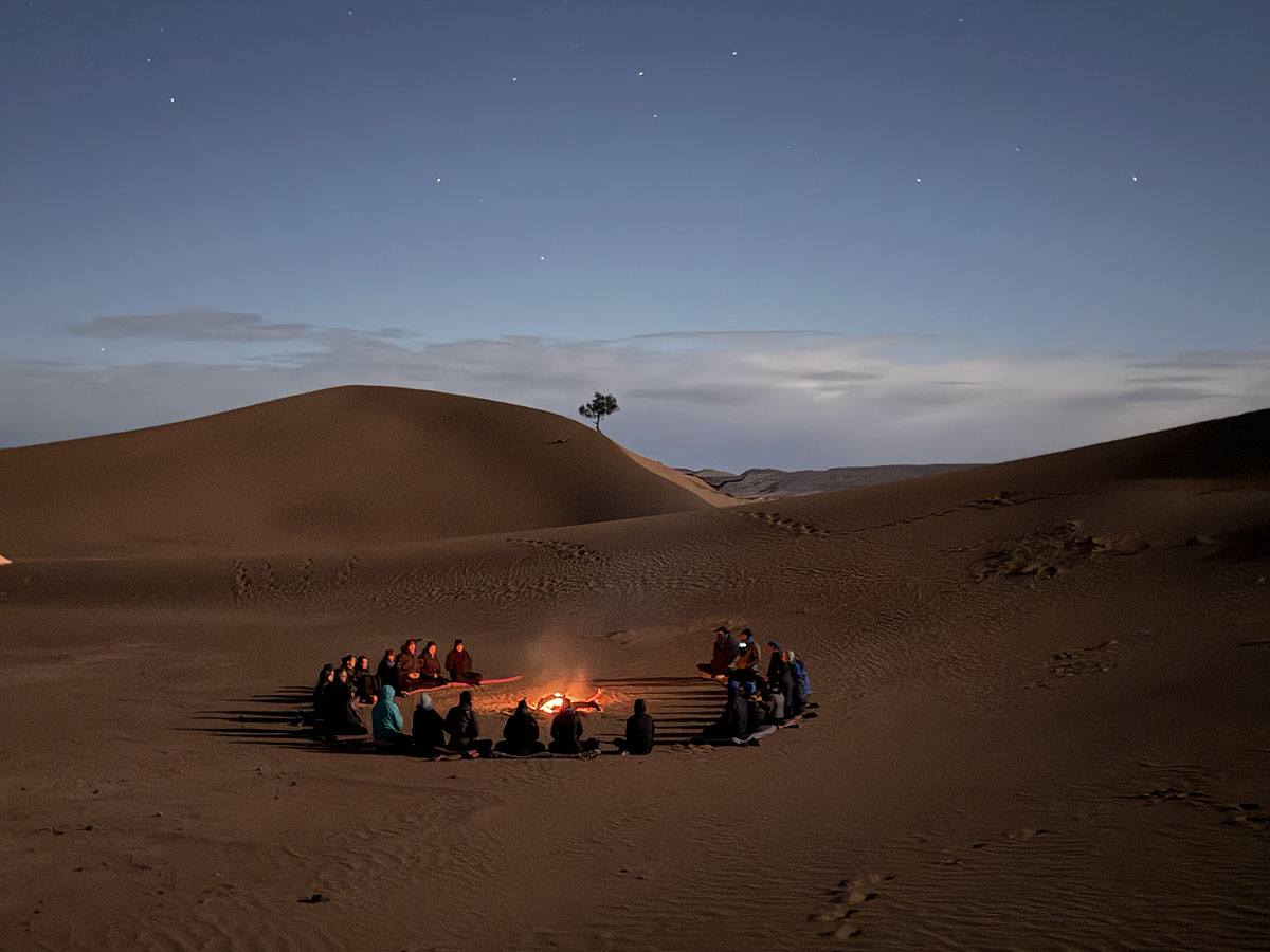 een groep mensen zit in het donker in een cirkel rond een kampvuur in de woestijn